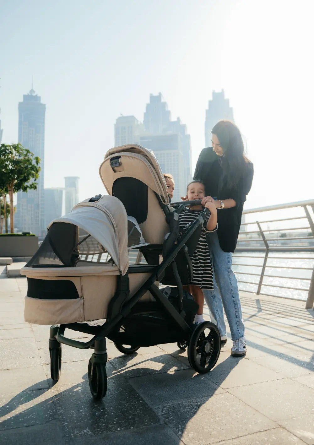 Eine Frau ist mit drei Kindern an einer Uferpromenade in der Stadt unterwegs. Im Bild: das Kinderwagen-Set UPPAbaby 'Vista 3' Liam mit Babywanne, Sportsitz und Mitfahrbrett