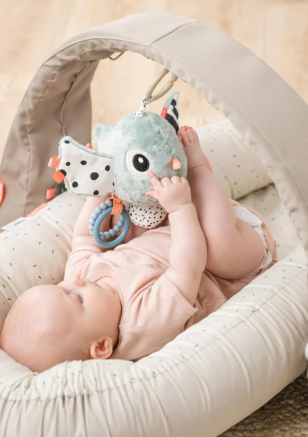 Ein im Nestchen liegendes Baby spielt mit dem Done by Deer Spielzeuganhänger 'Birdee' blau, der n einem Spielbogen befestigt ist.