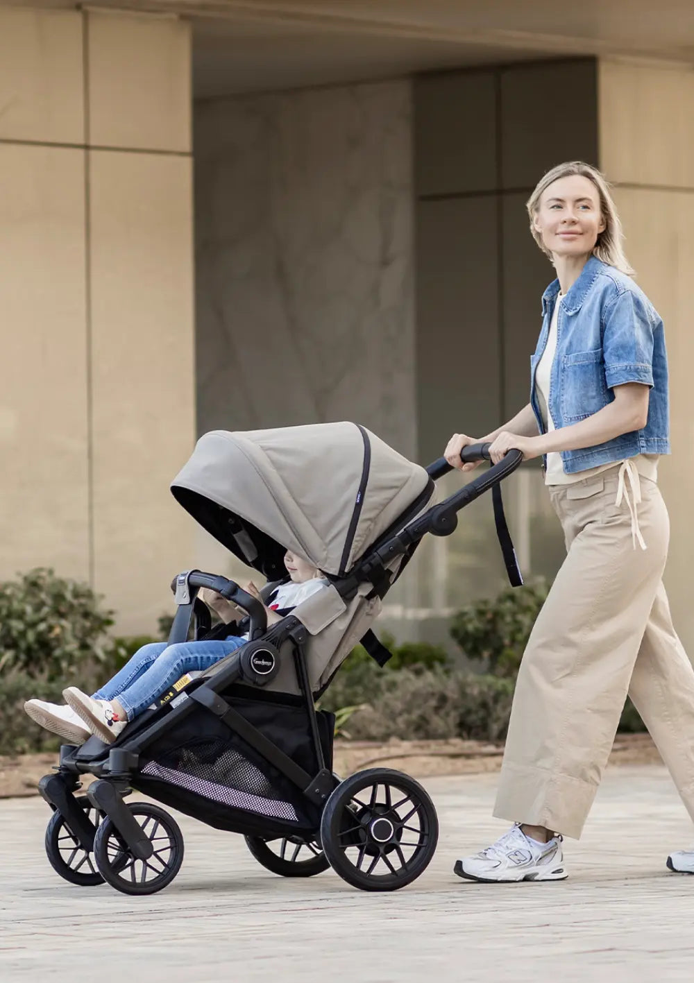 Eine Frau im Jeanshemd schiebt den beigefarbenen Emmaljunga Kinderwagen Sonic Stroller Urban Dune auf einem gepflasterten Platz.