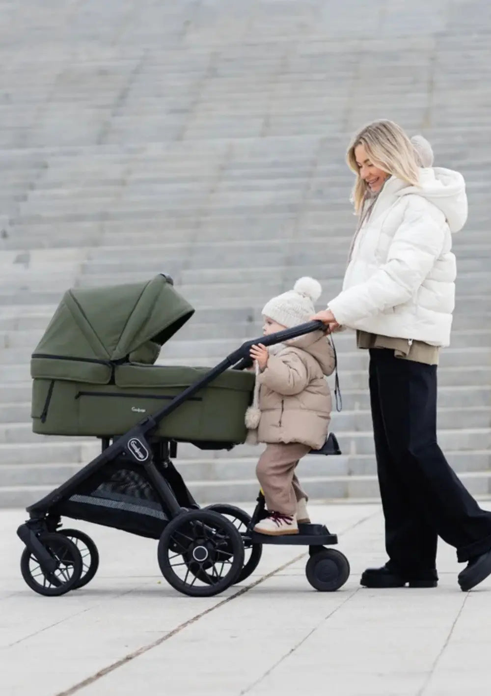 Eine Frau in einer weißen Daunenjacke schiebt einen Emmaljunga Kinderwagen, während ein Kleinkind auf dem befestigten Mitfahrbrett Roadeo Black mitfährt.
