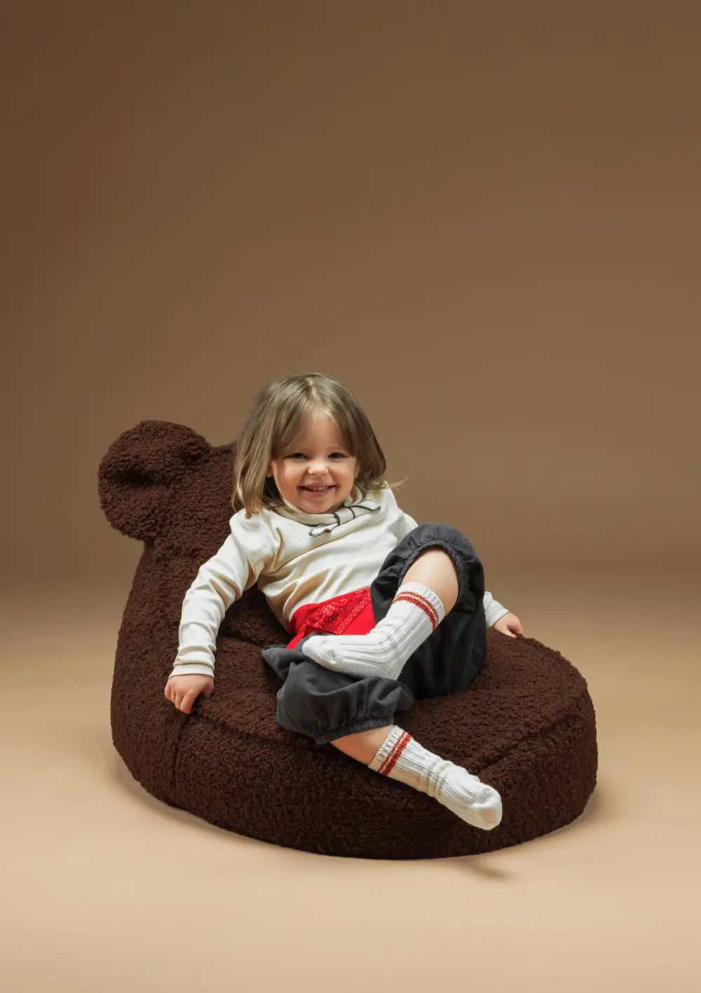 Ein fröhliches Kind sitzt gemütlich im braunen Wigiwama Bären Sitzsack Teddy Warm Hazel aus weichem Teddyplüsch mit niedlichen Bärenohren.
