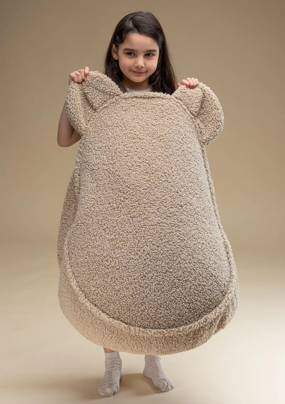 Ein Mädchen hält den beigefarbenen Wigiwama Bären Sitzsack Teddy Biscuit aufrecht vor sich, wodurch die Form mit den Bärenohren voll zur Geltung kommt.