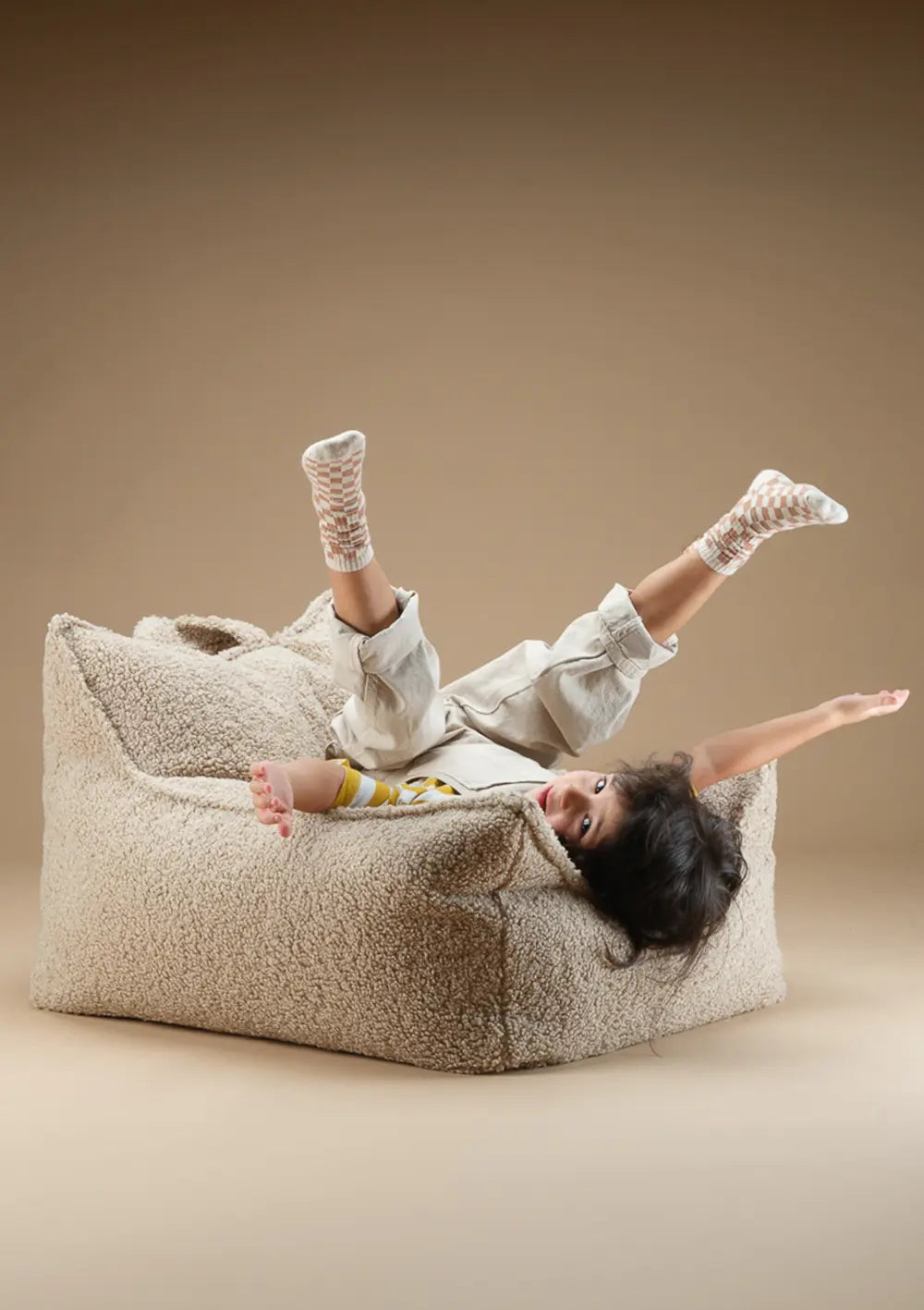 Ein Kind liegt fröhlich kopfüber im beigefarbenen Wigiwama Sitzsack Teddy Biscuit aus weichem Teddy-Plüsch und streckt Arme und Beine von sich.