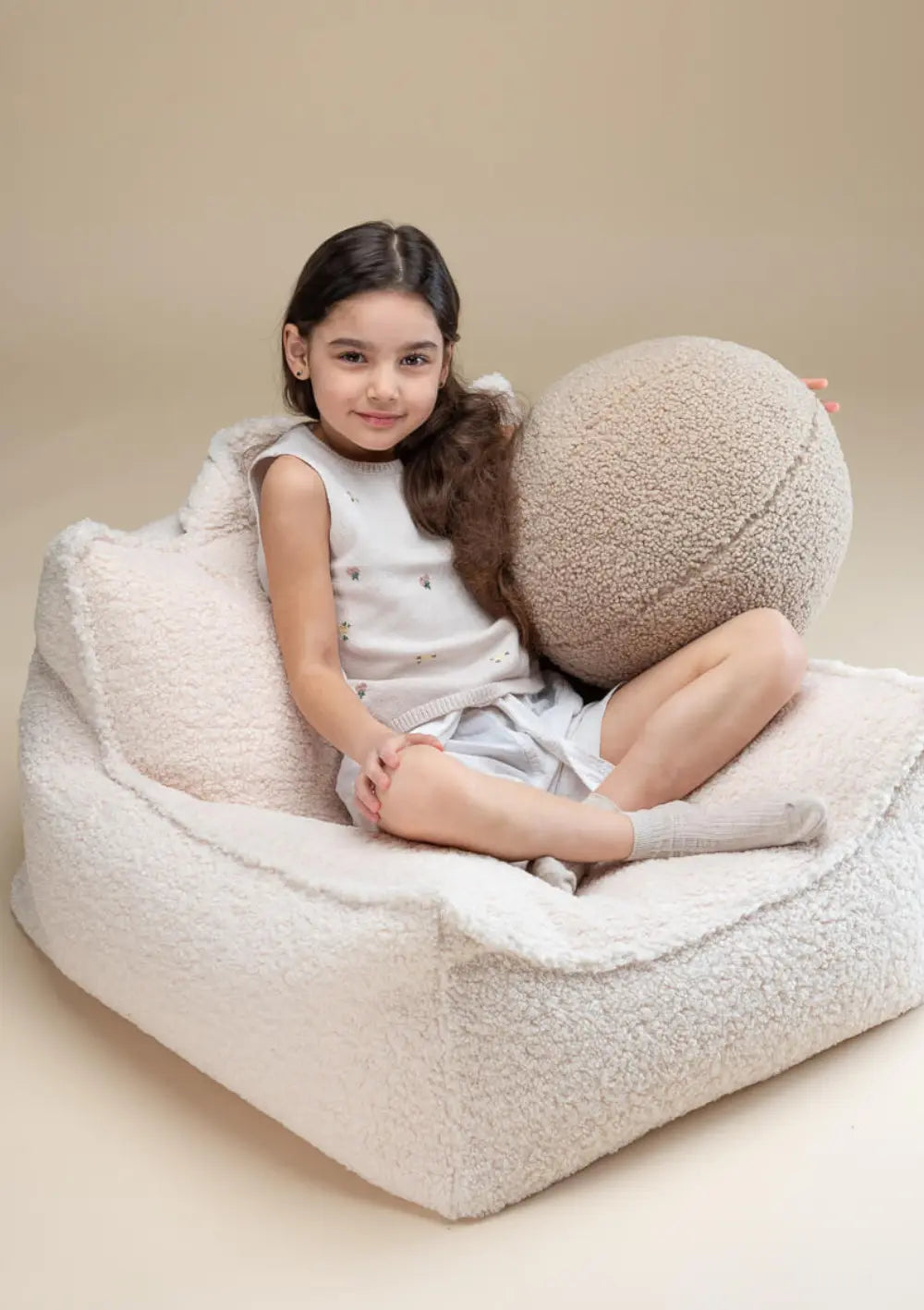 Ein Mädchen sitzt gemütlich im Schneidersitz mit einem runden Plüschkissen im cremeweißen Wigiwama Sitzsack Teddy Cream White aus weichem Teddystoff.