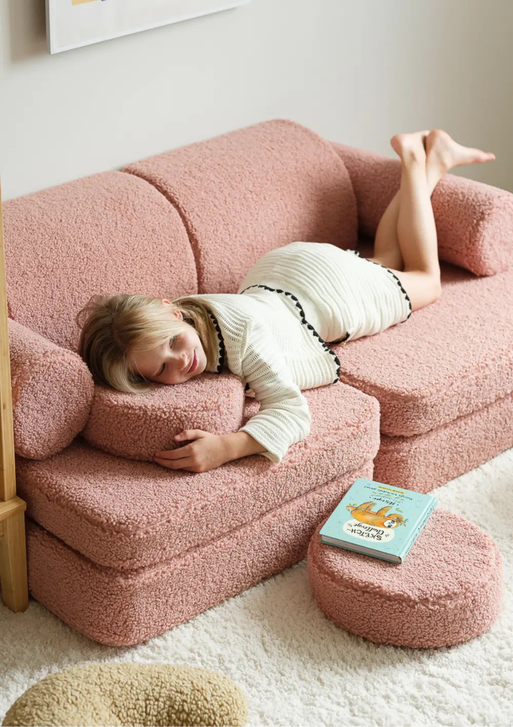 Ein Mädchen liegt gemütlich bauchlings auf dem rosafarbenen modularen Wigiwama Kindersofa Teddy Guava aus flauschigem Plüsch.