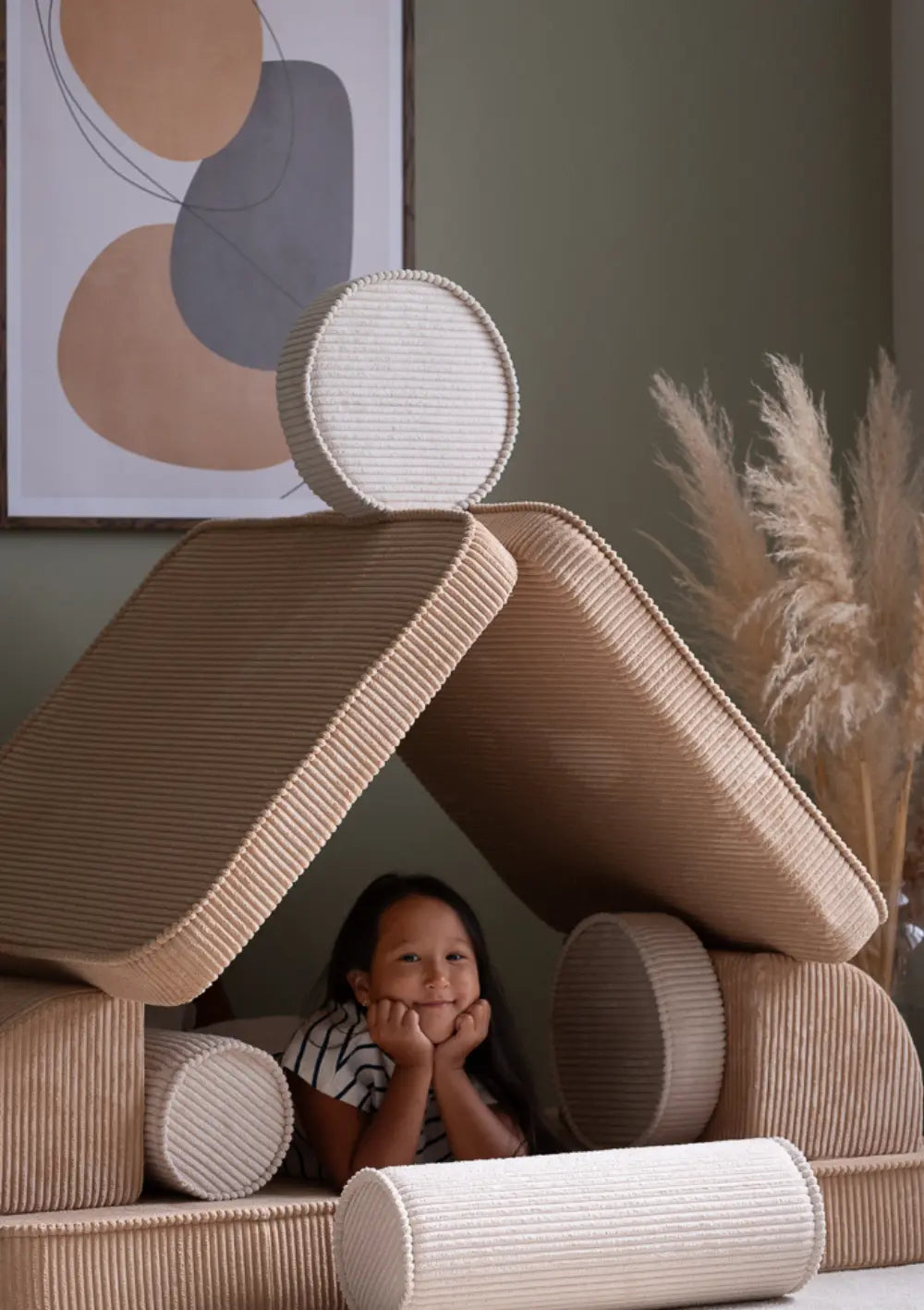 Ein Mädchen schaut lächelnd aus einer aus den Elementen des hellbraunen modularen Wigiwama Kindersofas Cord Brown Sugar gebauten Spielhöhle.