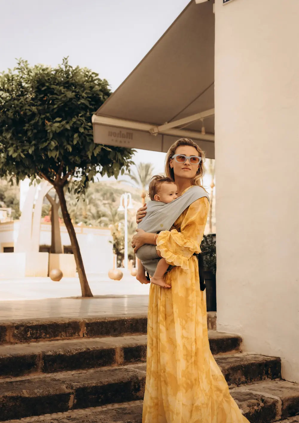 Eine Mutter im gelben Sommerkleid hält ihr Baby liebevoll im Arm, das sicher in der hellblauen Wildride Babytrage Firstride Bowie vor ihrer Brust getragen wird.