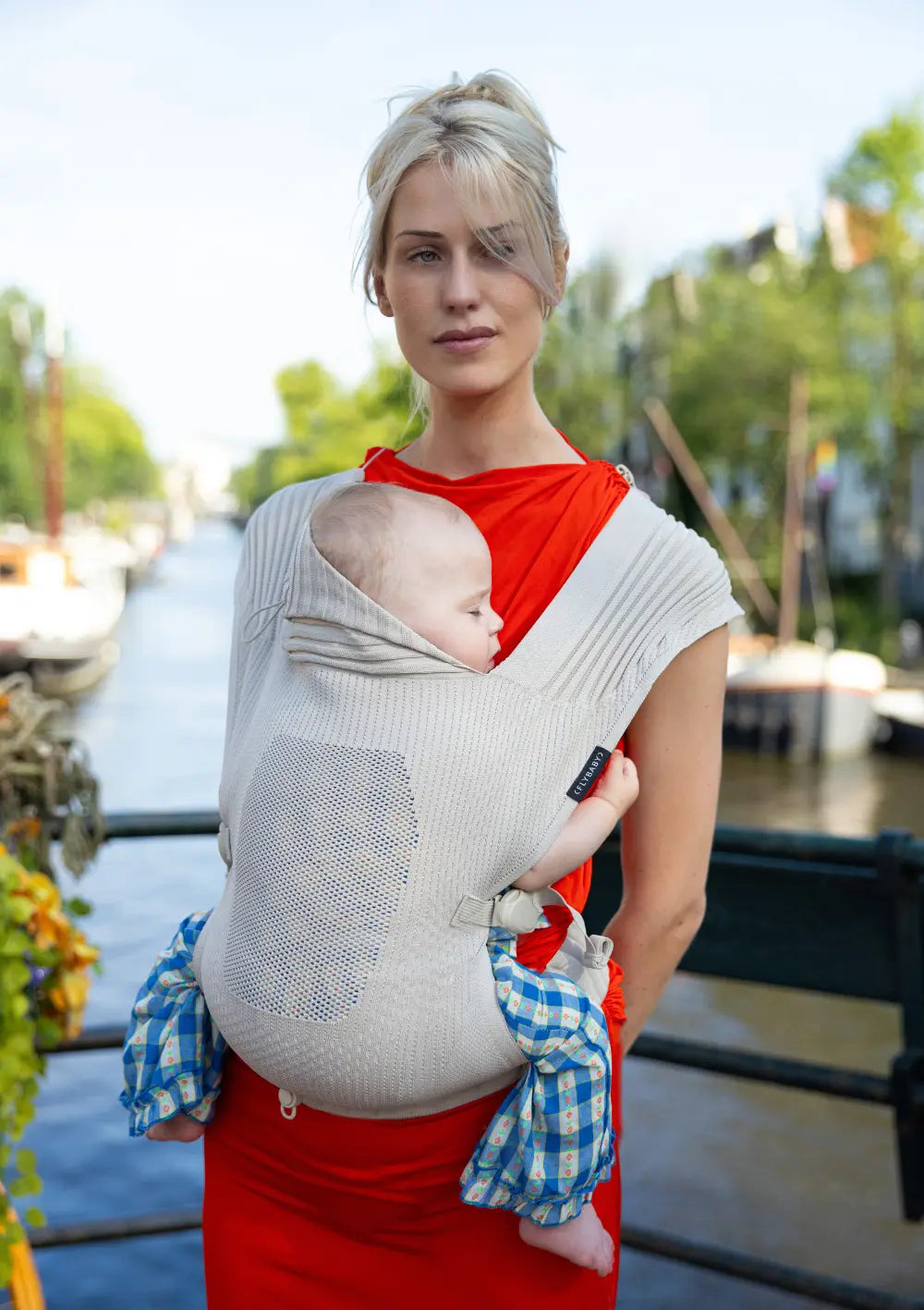 Eine Frau in einem roten Kleid trägt ein schlafendes Baby in der beige-cremefarbenen Flybaby Babytrage All-Cream vor einer Flusskulisse.