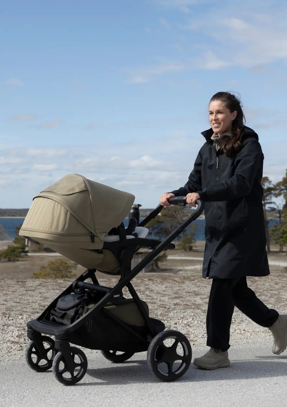 Eine Frau schiebt das Thule Kinderwagen 'Charm' Faded Khaki  mit Sportsitz auf einem befestigten Weg in einer Küstenlandschaft unter blauem Himmel.
