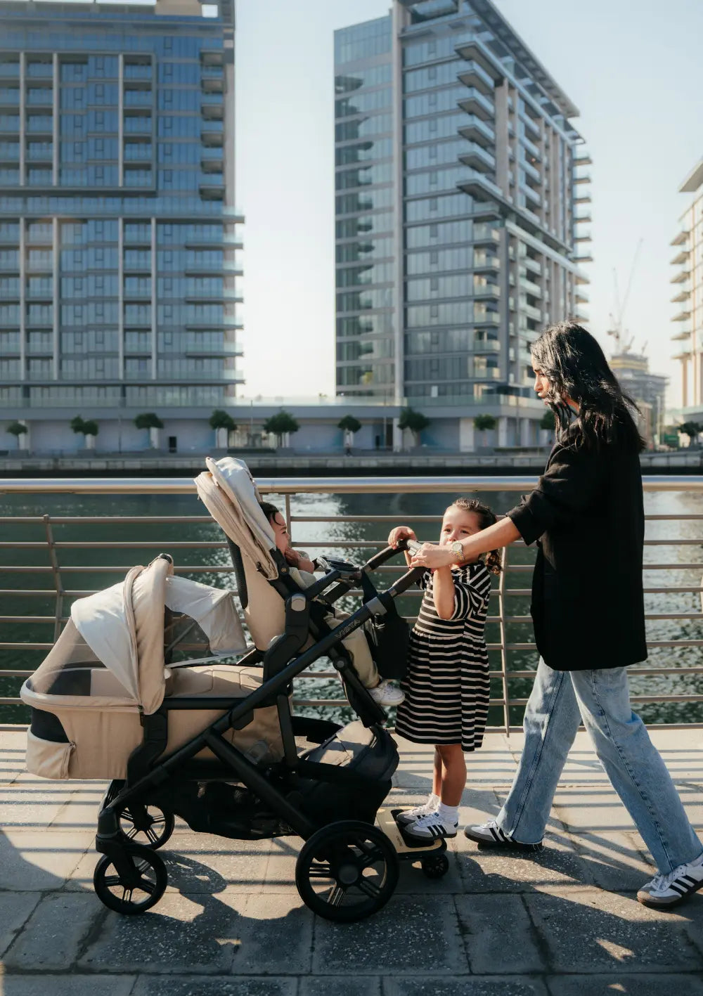 Eine Frau ist mit drei Kindern an einer Uferpromenade in der Stadt unterwegs. Im Bild: das Kinderwagen-Set UPPAbaby 'Vista 3' Liam mit Babywanne,   Sportsitz und Mitfahrbrett