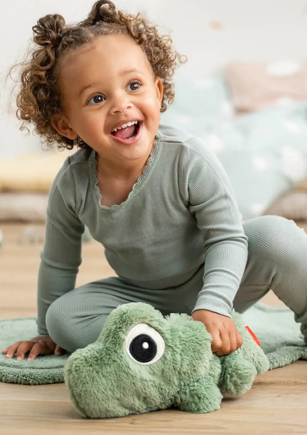 Ein fröhlich lachendes Kleinkind mit Locken kniet auf einer weichen Spielmatte und greift spielerisch nach dem grünen, plüschigen Krokodil Done by Deer Kuschelfreund Croco Grün mit den typischen großen Kulleraugen.