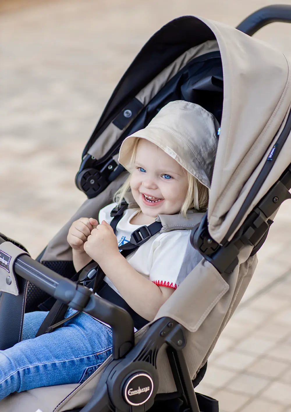 Nahaufnahme eines fröhlich lachenden Kleinkinds mit Sonnenhut, das sicher angeschnallt im beigefarbenen Emmaljunga Kinderwagen Sonic Stroller Urban Dune sitzt.