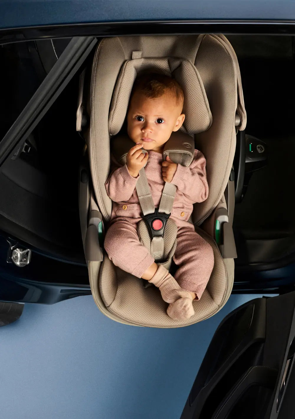 Draufsicht auf ein Baby, das in einem rosa Strampler und Söckchen in der  Maxi-Cosi Pebble Slide Pro Babyschale liegt und direkt in die Kamera blickt; der 3-Punkt-Gurt ist korrekt geschlossen und die seitlichen Gurtführungen sind sichtbar.