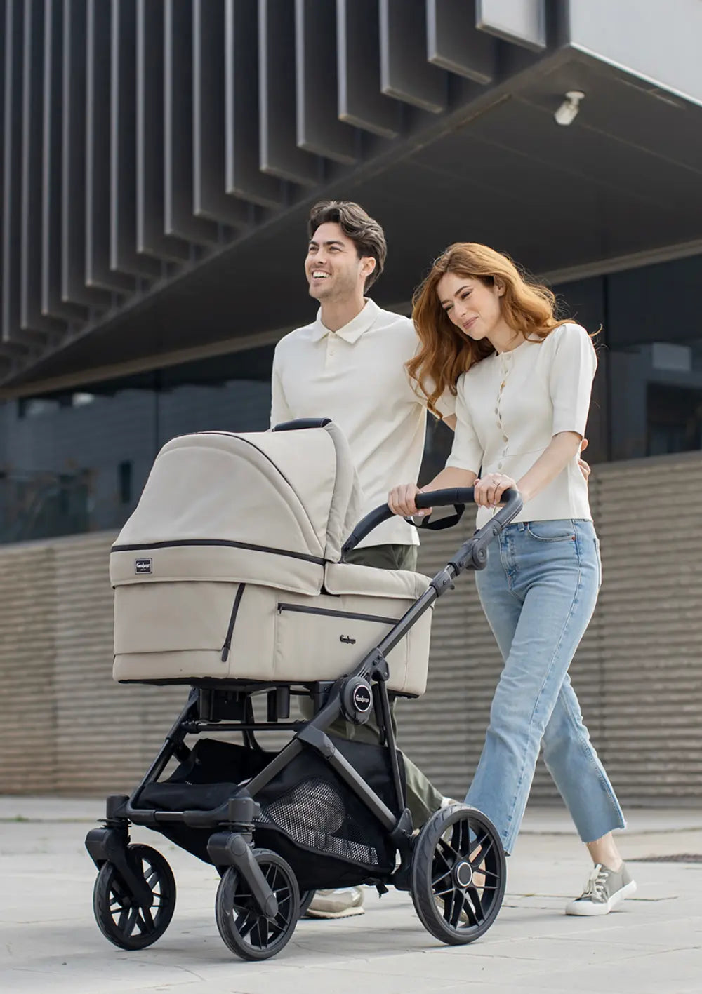 Ein Paar spaziert lächelnd vor moderner Architektur und schiebt den beigefarbenen Kombikinderwagen mit Babywanne: Emmaljunga Kinderwagen 'SUPER SONIC' Urban Dune