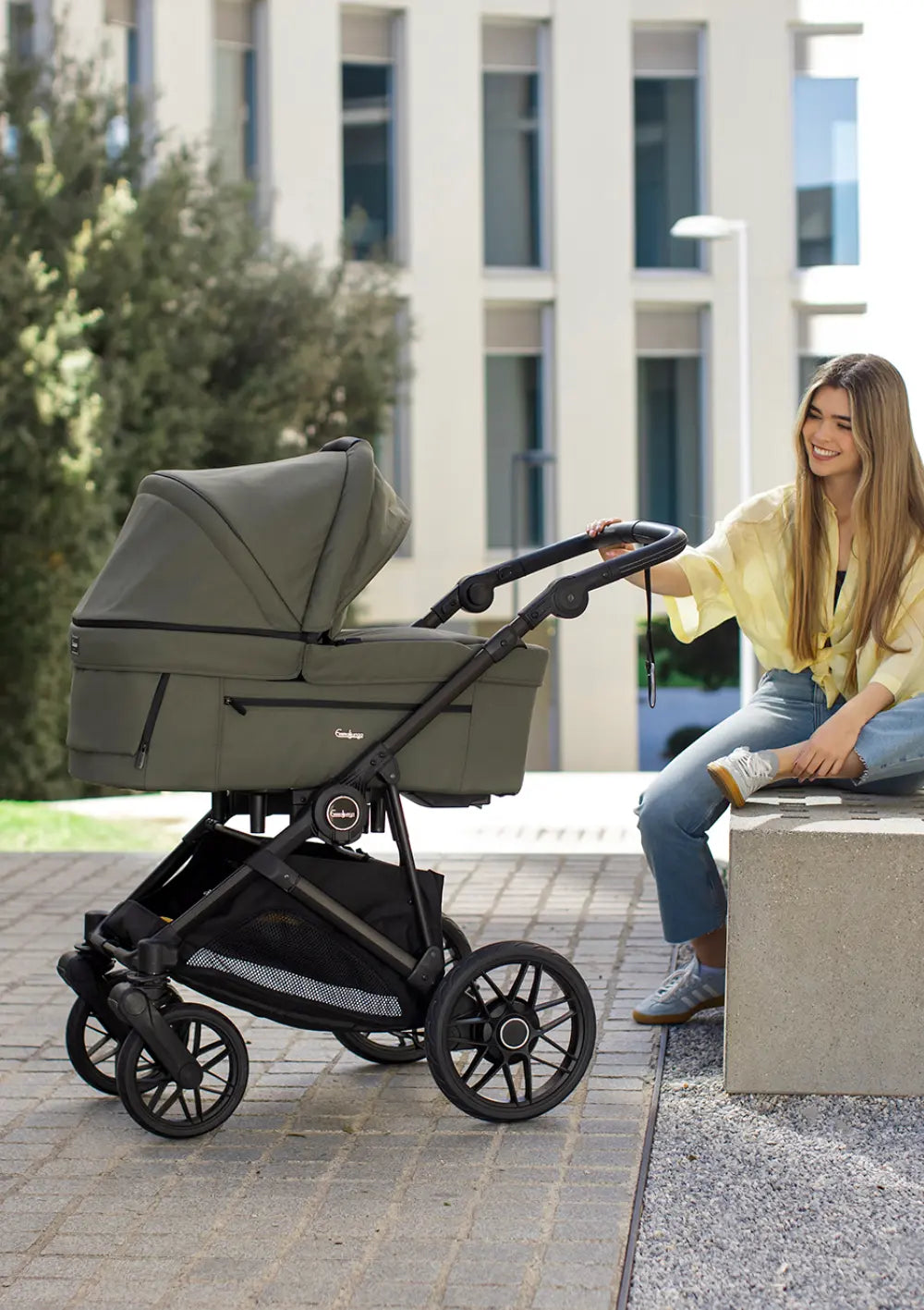 Eine Frau sitzt lächelnd auf einer Steinbank und hält den Griff des olivgrünen Kombikinderwagens mit Babywanne: Emmaljunga Kinderwagen 'SUPER SONIC' Urban Green.