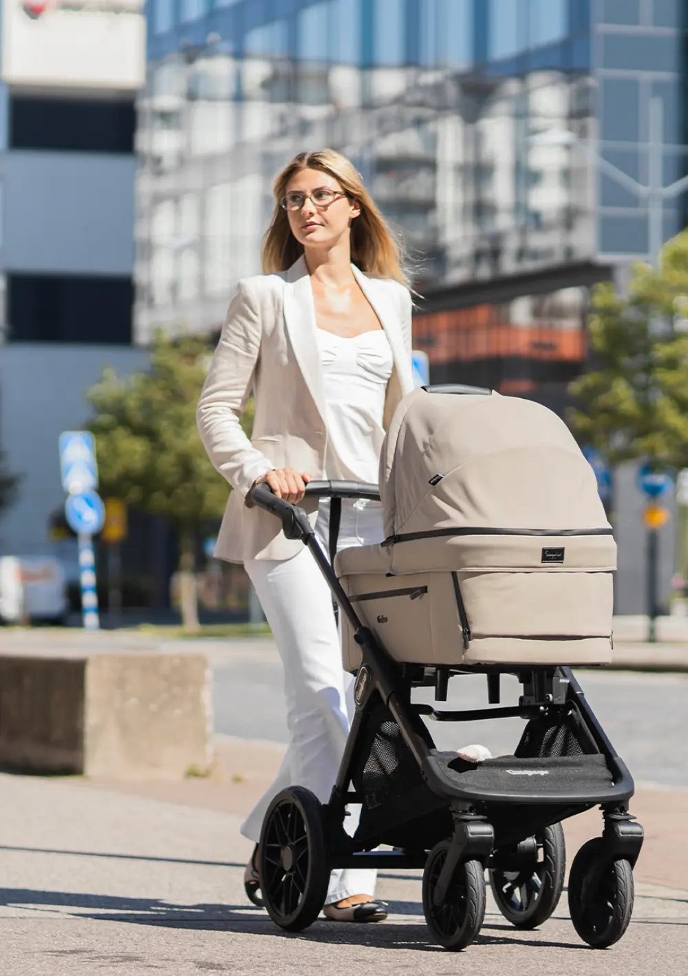 Eine Frau im hellen Blazer schiebt den beigefarbenen Kombikinderwagen mit Babywanne Emmaljunga SENTO PRO Urban Dune auf einem Gehweg im urbanen Umfeld.