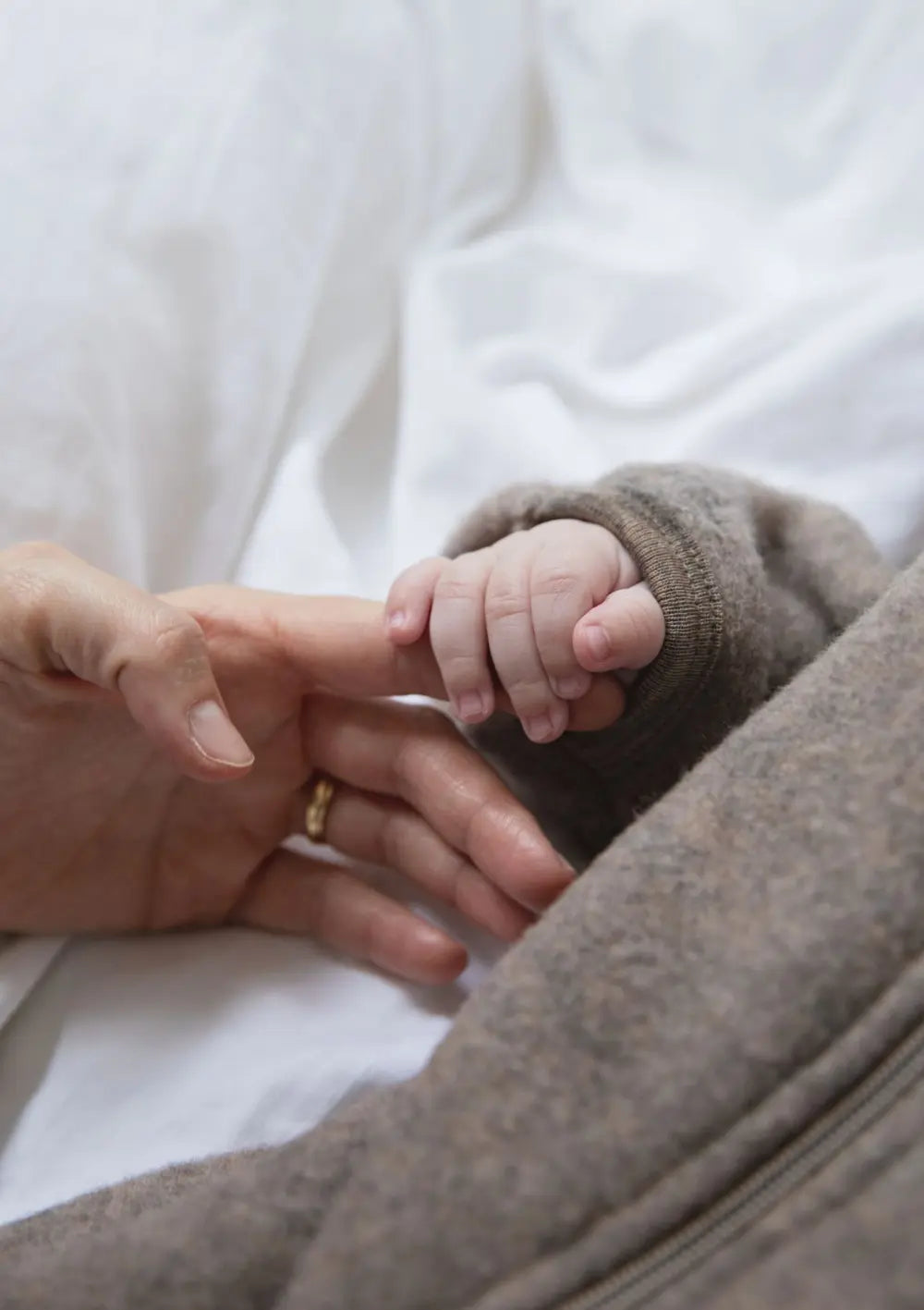 Nahaufnahme der Hände von Mama und Baby; das Baby trägt den Engel Wollfleece Zip Overall mit Kapuze und liegt auf dem Bett