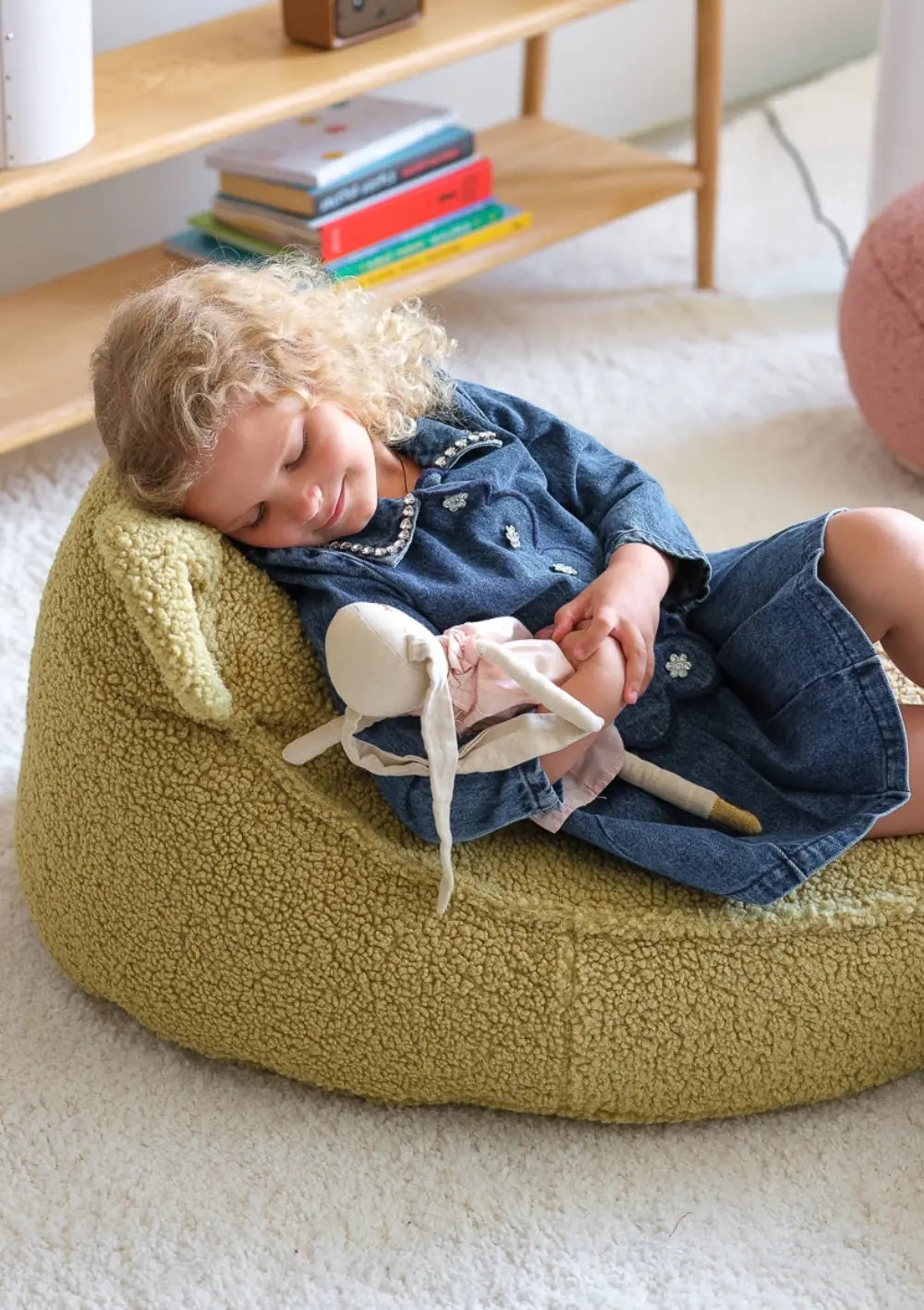 Ein kleines Mädchen mit blonden Locken kuschelt im hellgrünen Wigiwama Bären Sitzsack Teddy Matcha mit einer Stoffpuppe im Arm.