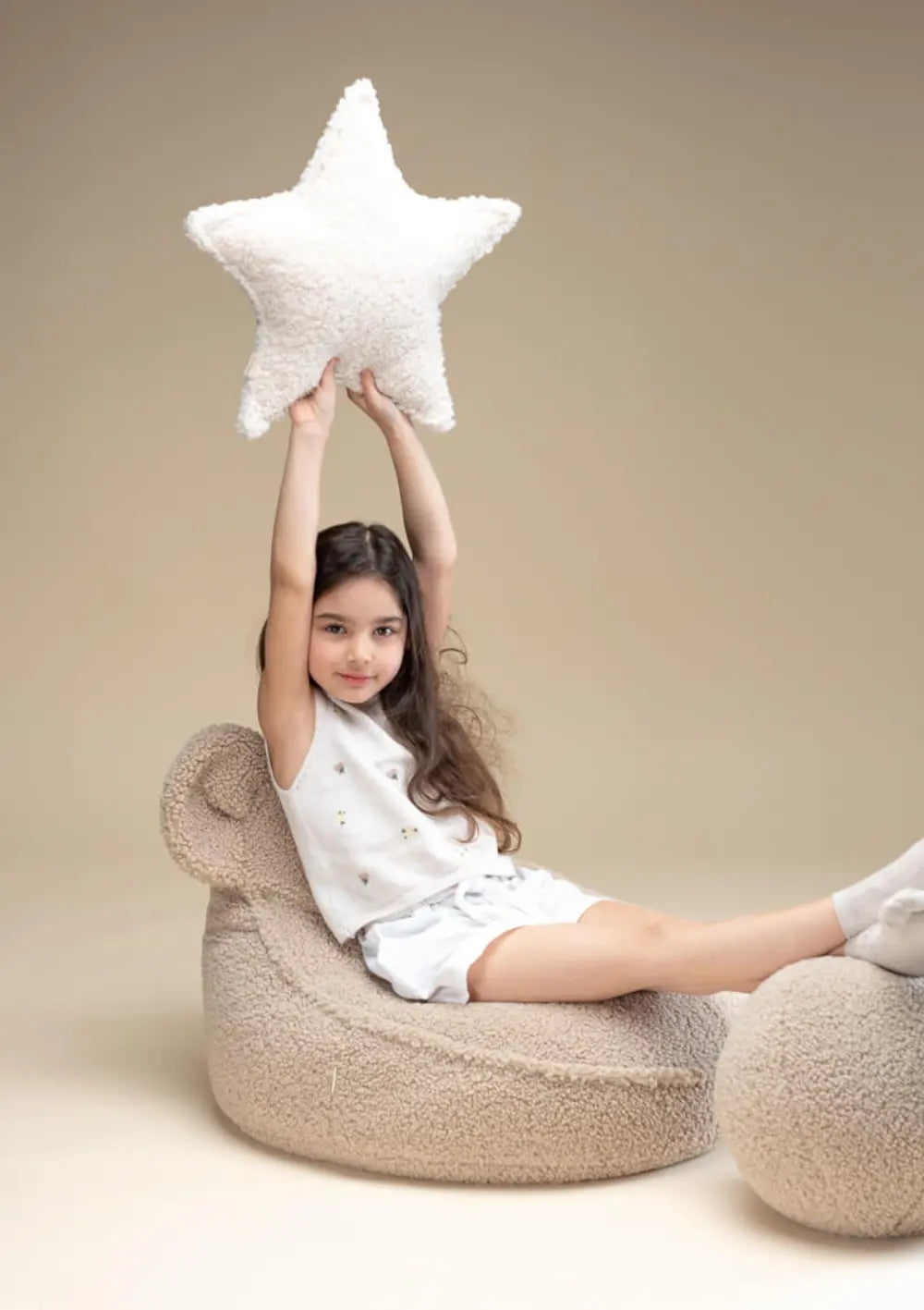 Ein Mädchen sitzt entspannt im beigefarbenen Wigiwama Bären Sitzsack Teddy Biscuit, legt die Füße auf einem passenden Pouf ab und hält ein sternförmiges Kissen hoch.