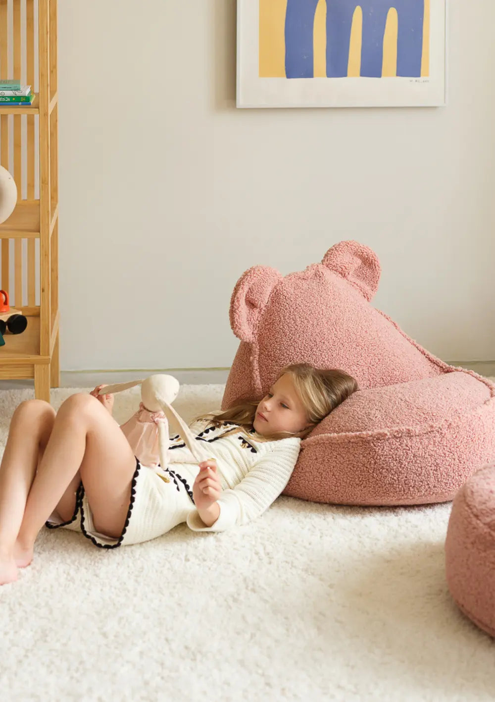 Ein Mädchen liegt entspannt auf einem hellen Teppich und lehnt mit dem Kopf am altrosa Wigiwama Bären Sitzsack Teddy Guava, während sie mit einem Stoffhasen spielt