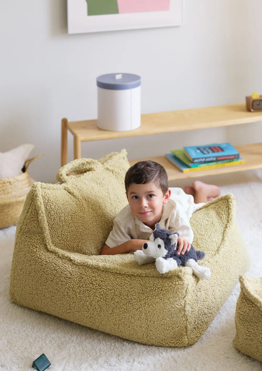 Ein Junge liegt bauchlings mit einem Kuschelhund im olivegrünen Wigiwama Sitzsack Teddy Matcha aus weichem Plüsch und blickt lächelnd in die Kamera.