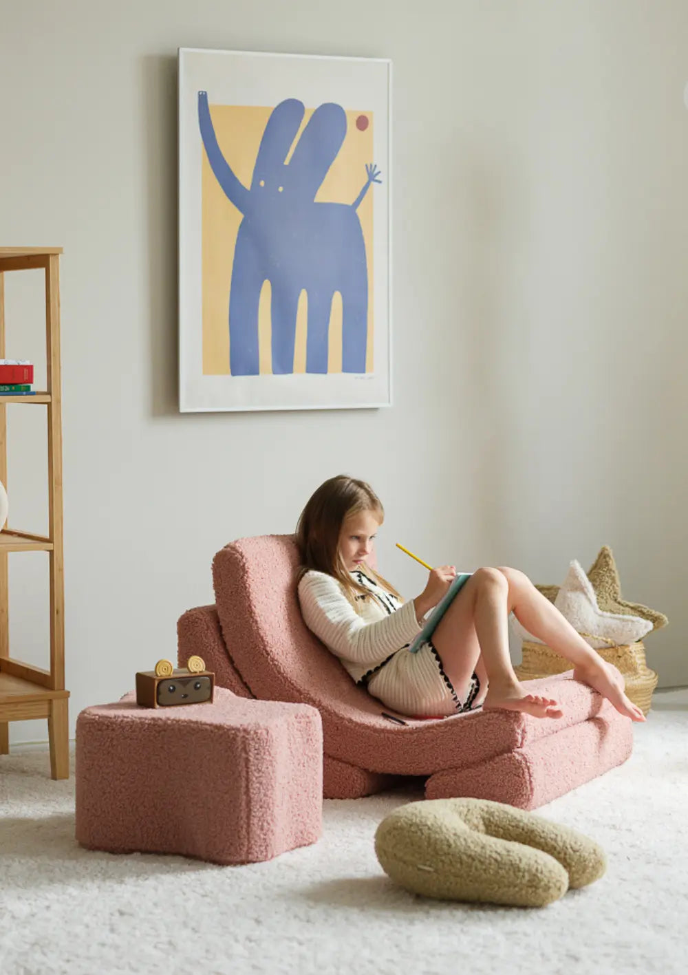 Ein Mädchen sitzt gemütlich im altrosafarbenen Wigiwama Kindersessel Moon Chair Teddy Guava und zeichnet konzentriert in einen Block.