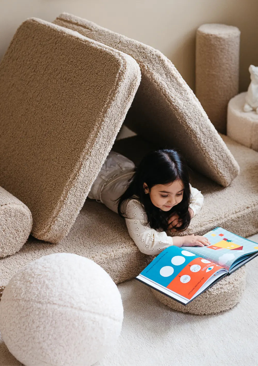 Ein Mädchen liegt gemütlich in einer aus den Elementen des beigefarbenen modularen Wigiwama Kindersofas Teddy Biscuit gebauten Höhle und liest ein Buch.