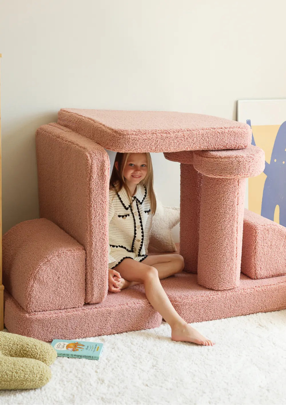 Ein Mädchen sitzt in einem aus den Bausteinen des rosafarbenen modularen Wigiwama Kindersofas Teddy Guava gebauten Spielhaus.