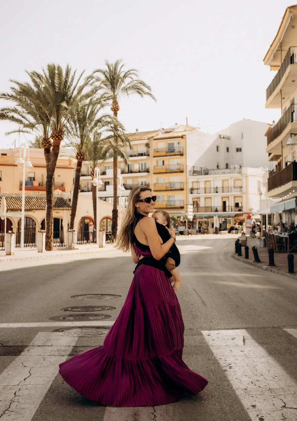 Eine lächelnde Frau im magenta Plissee-Kleid läuft über die Straße und trägt ihr Baby vor der Brust in der Wildride Babytrage Firstride Bowie Minimal Schwarz.
