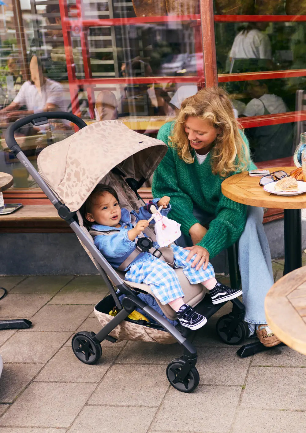 Eine lächelnde Frau sitzt in einem Café neben dem Joolz Buggy Aer2 Chic Renaissance Sandy Taupe, in dem ein Kind mit einer Puppe spielt.